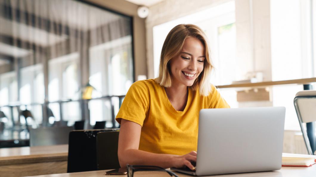 Person in a yellow shirt working on a laptop at a table in a bright, modern cafe setting.
